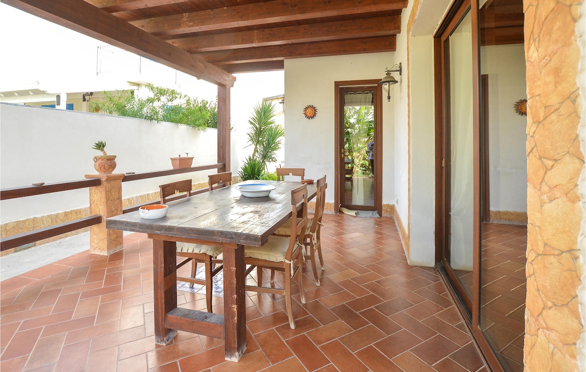 An outdoor terrace is shown, featuring a large wooden dining table surrounded by several chairs. The space is covered by a wooden roof, with tiled flooring and potted plants enhancing the relaxing atmosphere. Large glass doors provide access to an indoor area.