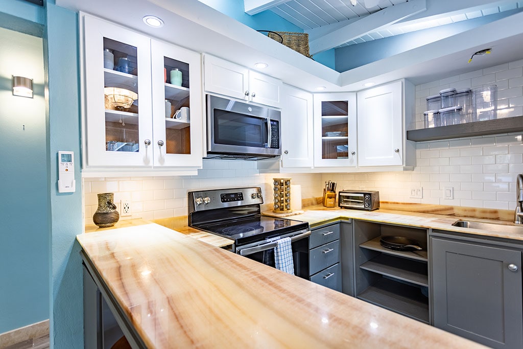 A modern kitchen features light-colored cabinetry and a stone countertop. Stainless steel appliances, including a microwave and oven, are incorporated into the design. Glass-fronted shelves display dishware, while under-cabinet lighting enhances the workspace's functionality.