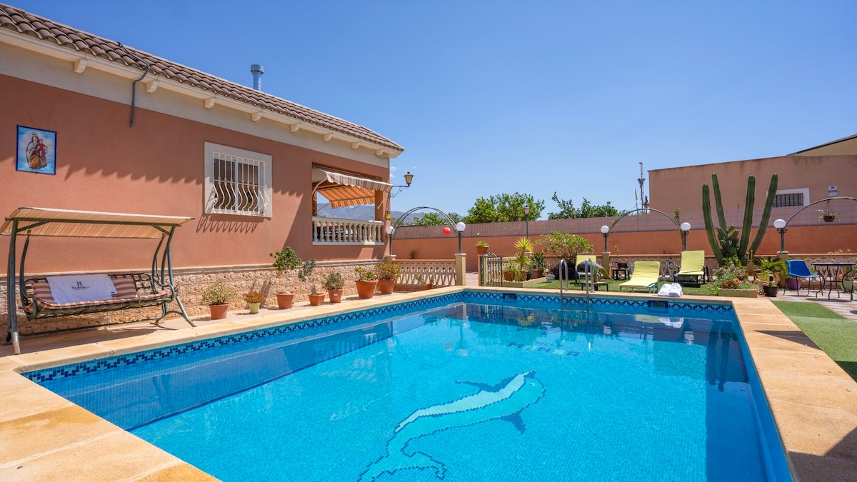 A private pool is featured in the foreground, showcasing a tiled design with a dolphin motif. Surrounding the pool, potted plants enhance the outdoor space. A swing seat is positioned nearby, with sun loungers visible in a sunlit garden area.