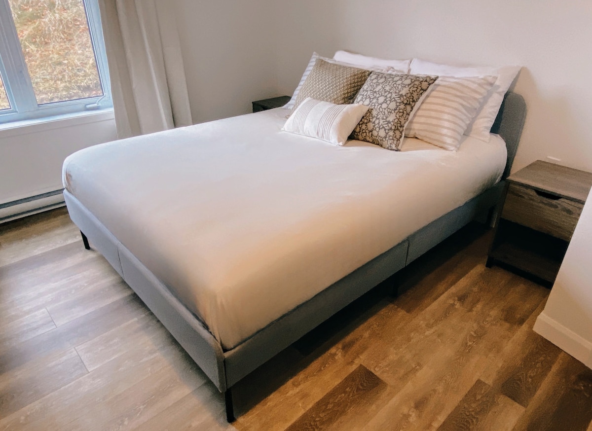 A spacious bed is dressed in crisp white linens, complemented by decorative pillows in neutral tones. Two nightstands are positioned beside the bed, adding functional decor. Natural light filters through a large window with sheer curtains, illuminating the warm wood flooring.