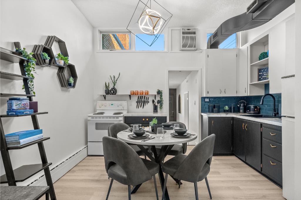 A modern kitchen area features a round dining table set for four, accompanied by stylish chairs. Black cabinetry contrasts with a bright blue backsplash. Open shelving adds decorative plants, while a geometric light fixture hangs above. Natural light enters through multiple windows, enhancing the space.