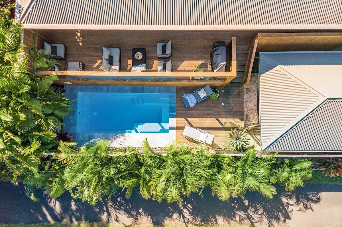 An aerial view captures a tranquil outdoor pool flanked by sun loungers and a shaded lounging area. Lush greenery surrounds the space, adding to the secluded atmosphere. The deck is lined with modern outdoor furniture, providing a welcoming spot for relaxation.