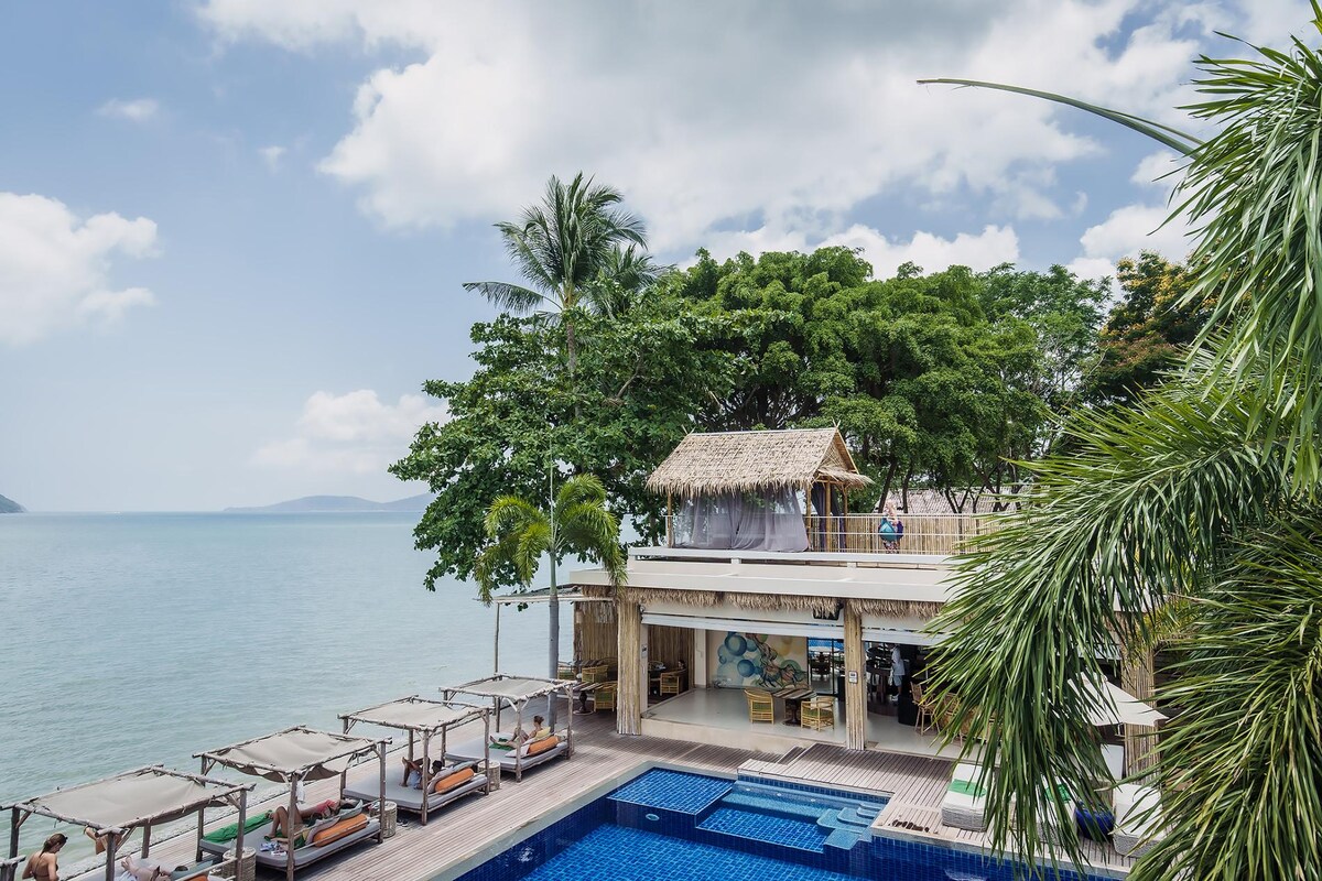 A serene beachfront scene showcases a multi-level structure with a thatched roof, nestled among lush trees. A sparkling blue pool reflects the sky, surrounded by lounge chairs. The calm waters of Chalong Bay are visible in the background, enhancing the tranquil atmosphere.