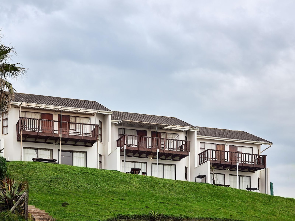 A three-story chalet is situated on a lush green hillside, featuring multiple balconies with wooden railings. The exterior is painted in neutral tones, complemented by large glass windows that provide views of the surrounding landscape.