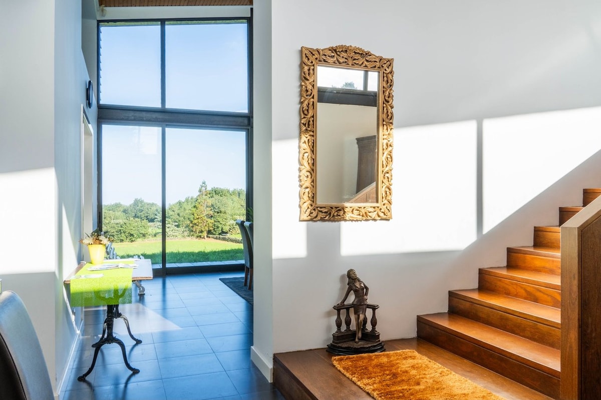 An open entryway showcases a wooden staircase leading to the upper level. A decorative mirror with an ornate frame is mounted on the wall, reflecting light from large glass doors that overlook a green landscape. A small table with a vibrant tablecloth is positioned nearby.