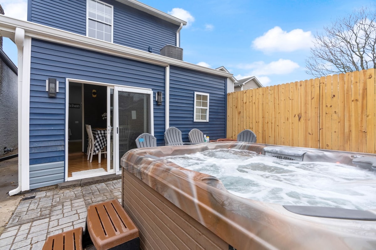 A private hot tub is set in the outdoor area, with bubbling water creating an inviting scene. Wooden seating is arranged nearby, and a cozy dining space can be glimpsed through the open door, surrounded by a wooden fence for privacy.