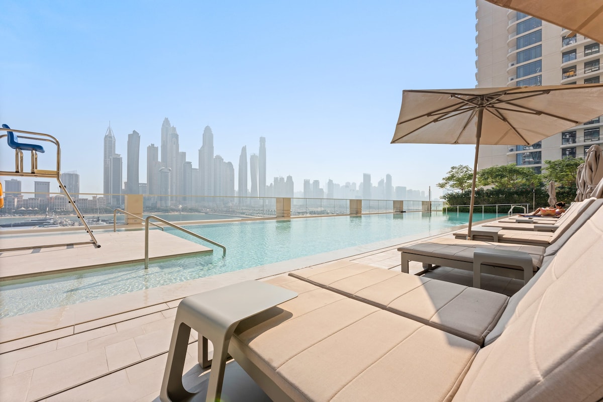 The outdoor pool area features comfortable lounge chairs arranged along the water's edge. A large umbrella provides shade, while the serene pool reflects the skyline of Dubai's modern towers in the background, creating a tranquil atmosphere for relaxation.