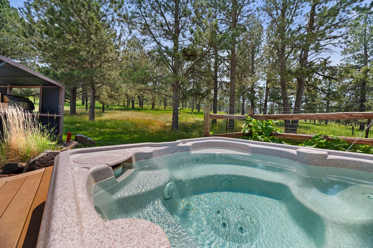 A hot tub is positioned on a wooden deck, surrounded by lush greenery and tall trees. The inviting water shows gentle ripples, reflecting the sunlight. A fenced area is visible in the background, enhancing the serene outdoor setting.