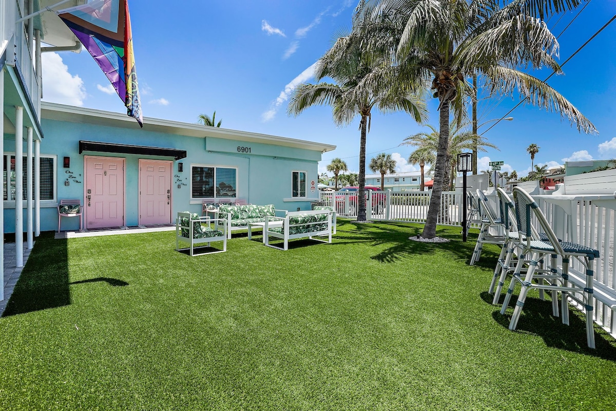 A vibrant courtyard is presented with artificial grass, bordered by palm trees. Comfortable outdoor seating with cushions is arranged around a coffee table, inviting relaxation. Two colorful doors are situated in the background, adding charm to the sunny atmosphere.