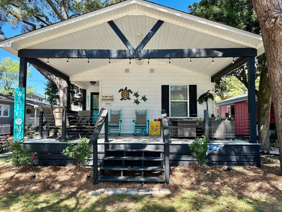 A charming front porch features a wooden railing and steps leading to the entrance. Two colorful rocking chairs sit under a covered area, accompanied by comfortable seating. Lush greenery and a welcome sign add to the inviting outdoor space.