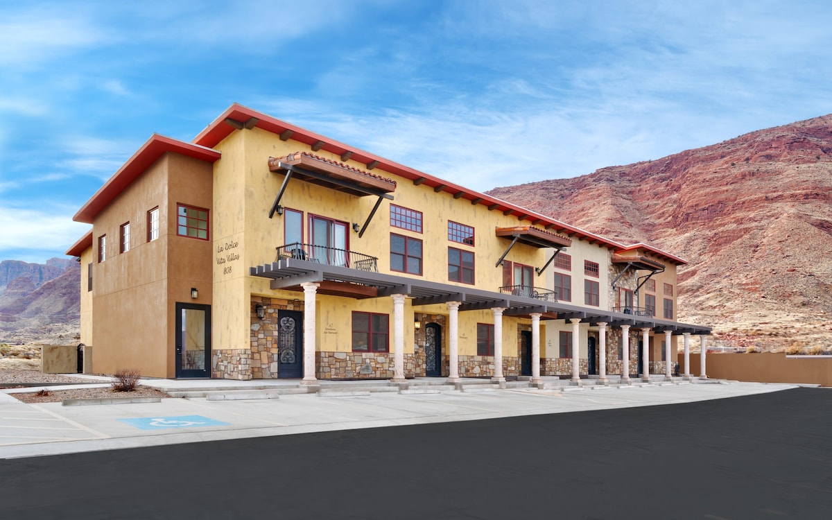 A modern, two-story building is depicted against a backdrop of red rock formations, showcasing a blend of earthy tones and architectural details. Multiple entrances with decorative ironwork are visible, along with designated parking spaces in front.