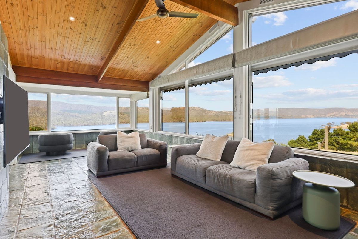 A spacious living area features large windows showcasing expansive views of Lake Eildon and the surrounding landscape. Comfortable grey sofas are positioned for relaxation, while natural wood beams highlight the high ceiling. A small round table stands nearby, complementing the tranquil setting.
