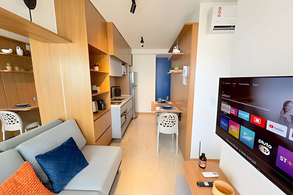 A modern living space is highlighted with wooden cabinetry and sleek appliances. A comfortable sofa is positioned near a smart TV. A dining area for two is visible, featuring minimalist design elements. Natural light brightens the space, creating a welcoming environment.