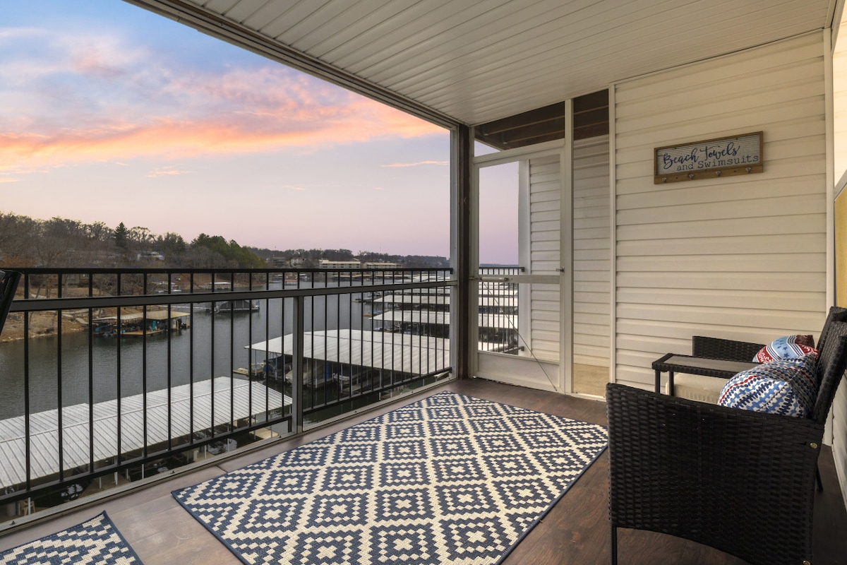 A screened-in balcony offers seating for two with black wicker chairs accented by colorful cushions. A decorative rug lies on the floor, complementing the view of the waterfront and marina beyond, illuminated by a soft twilight sky.