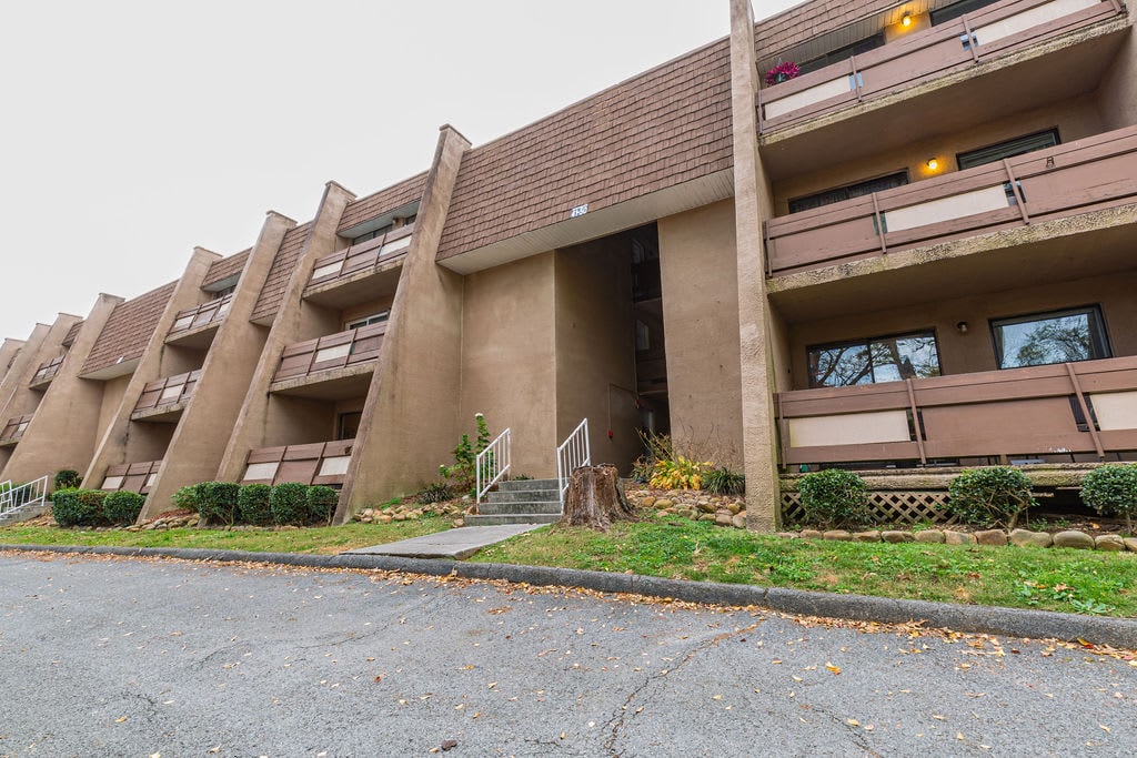 The exterior of the apartment complex features multiple two-story units. Staircase access is visible, leading to the entrance. Landscaped shrubs and small trees line the ground level, while light filters through the balconies, creating a welcoming façade.