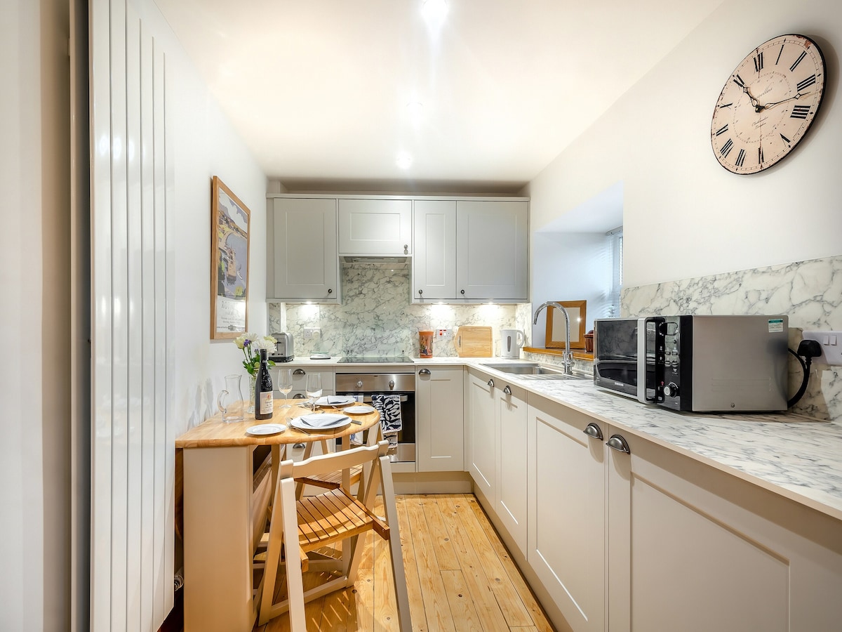 A well-equipped kitchen area features light-colored cabinetry, a marble countertop, and modern appliances including a microwave and oven. A small dining table made of wood provides seating for two, with a gentle hint of natural light illuminating the space.