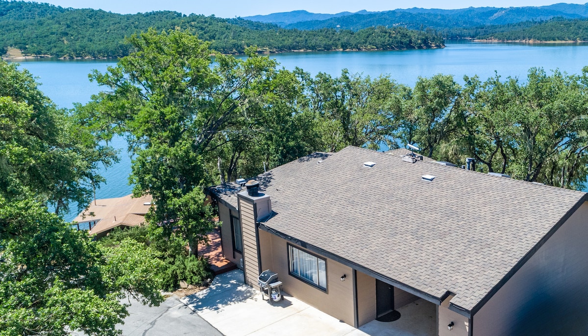 A spacious cabin is viewed from above, positioned along the shoreline of a calm lake. Surrounding trees provide greenery, and gentle hills are visible in the background against a clear blue sky.