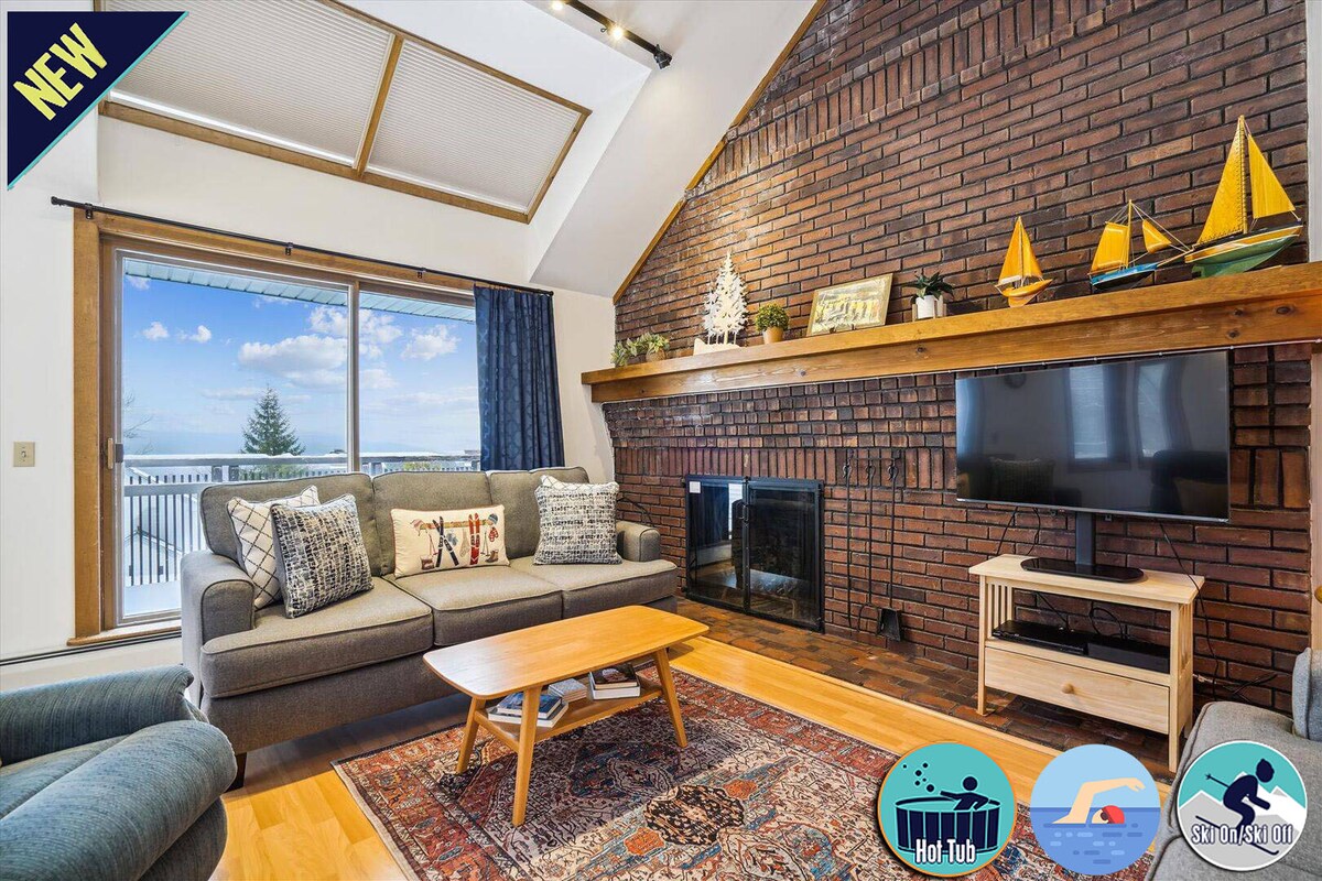 A cozy living room is presented with a brick wall and wood accents. A flat-screen TV is mounted on a stand, complemented by a wood-burning fireplace. Large windows provide natural light and views of the surrounding landscape, with a comfortable sofa and a coffee table enhancing the inviting space.