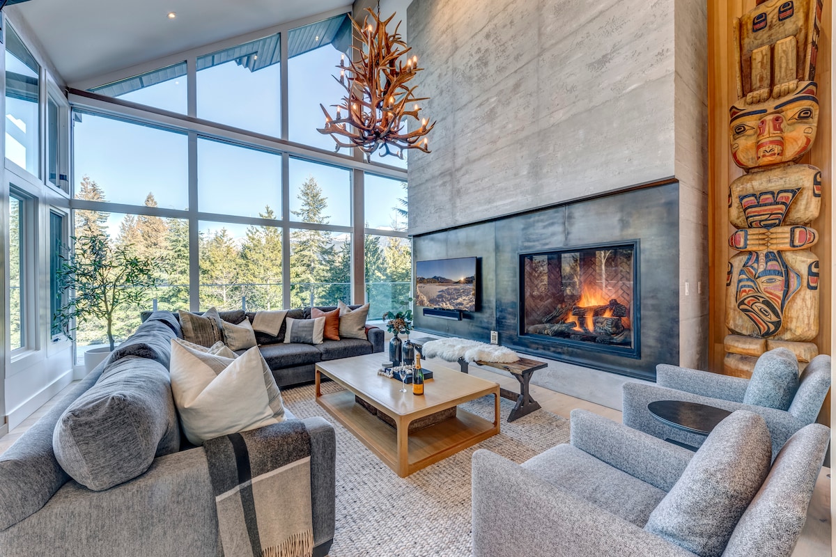 The spacious living area features large windows that fill the room with natural light, offering views of the surrounding landscape. A modern fireplace is centrally positioned, surrounded by comfortable seating arrangements. Textured artwork adorns the wall, and a wooden chandelier hangs above the seating area.