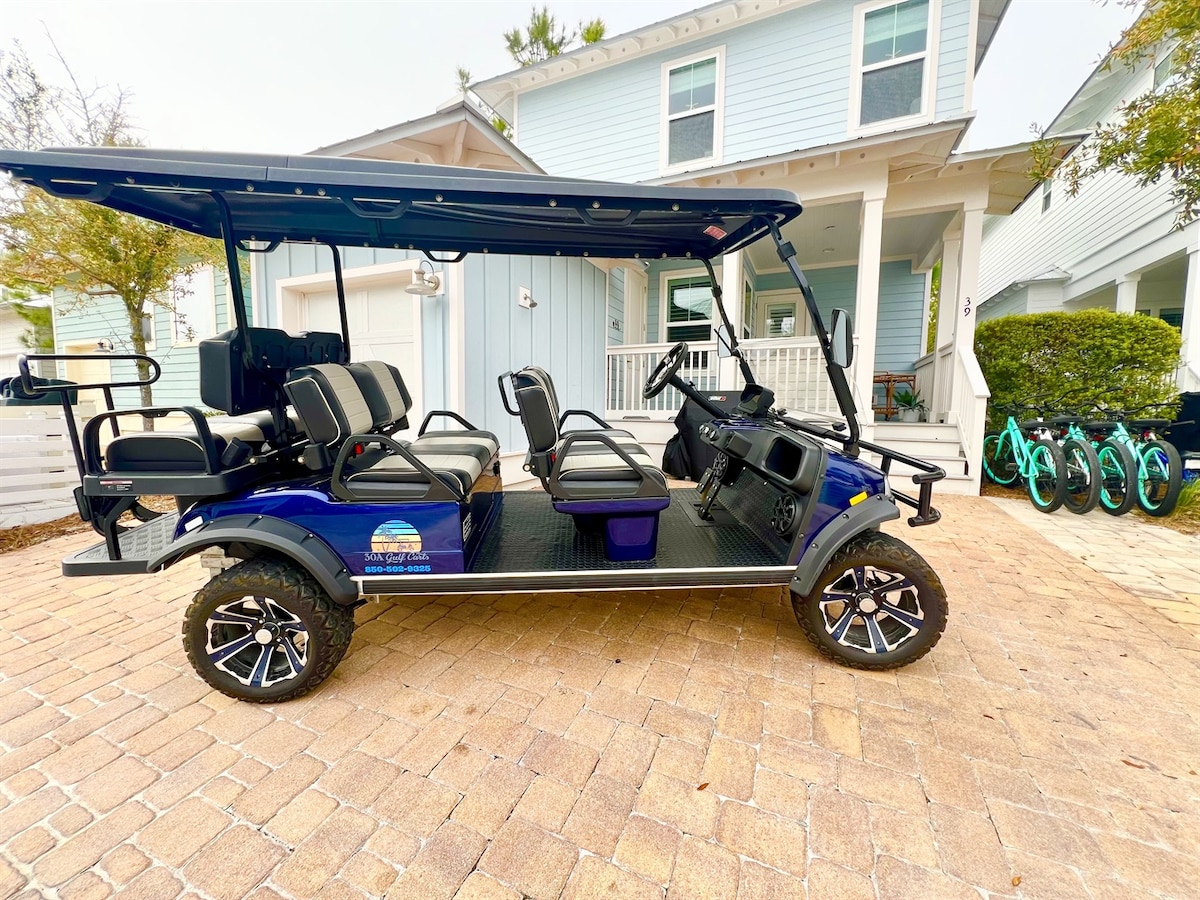 A six-seater golf cart is parked on the patterned brick driveway, showcasing a sleek blue exterior with comfortable seating. The background features a light blue house and several bicycles lined up to the side, indicating accessibility and mobility options.