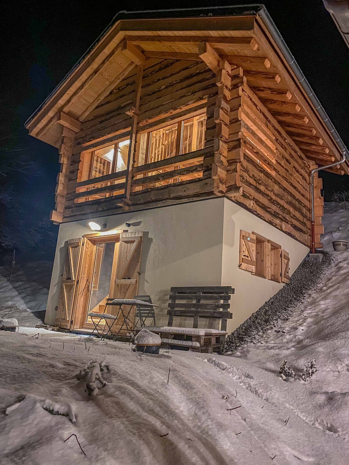 A charming two-story cabin features a wooden exterior with large windows allowing warm light to illuminate the snow-covered surroundings. A small outdoor seating area with two chairs and a table is visible, set against a backdrop of a peaceful winter landscape.