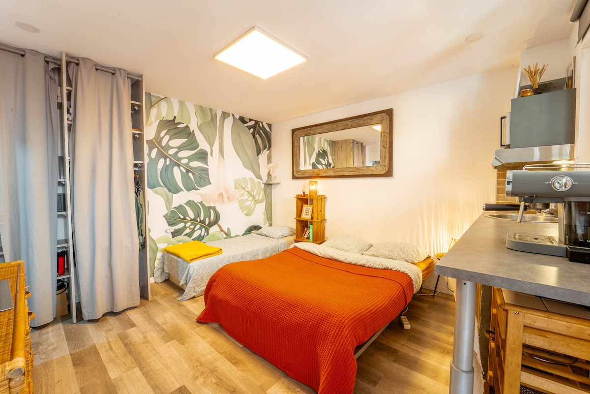 A welcoming sleeping area features a spacious bed covered with a warm orange blanket. A light tone wood floor complements the leafy wallpaper. A mirrored wall reflects natural light from the ceiling. A simple kitchenette with wooden shelves and a coffee maker is positioned along one wall.