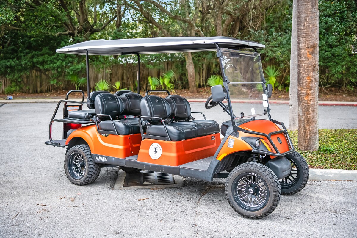 A six-seater golf cart is parked on a paved surface, featuring comfortable black seating and a canopy for shade. The cart's vibrant orange body is complemented by stylish alloy wheels. Lush greenery is visible in the background, enhancing the outdoor setting.