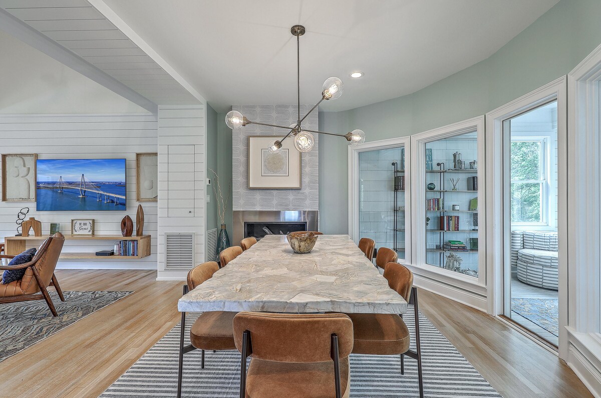 A spacious dining area is presented with a large stone dining table surrounded by eight upholstered chairs. Soft green walls and modern light fixtures enhance the elegant yet inviting space. Large windows invite natural light, highlighting the cozy seating area nearby.