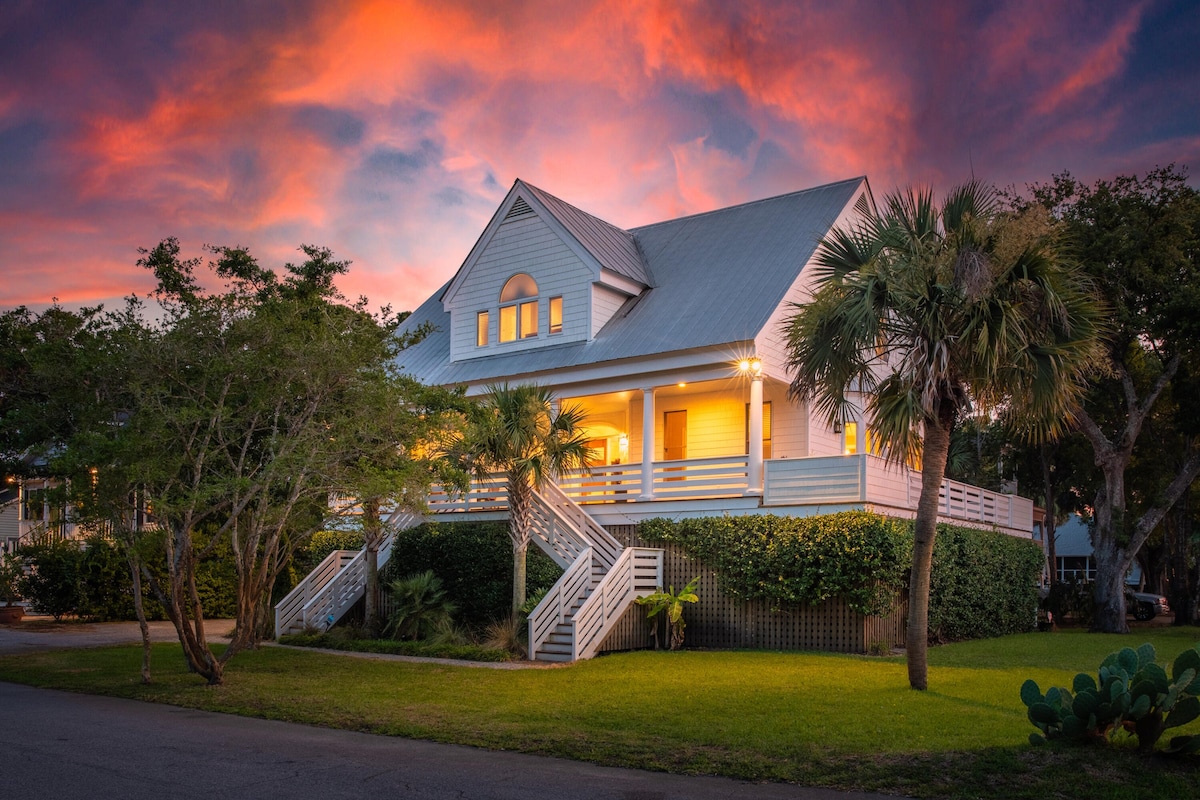 A coastal home is showcased against a vibrant sunset sky, featuring a wrap-around porch and multiple steps leading up to the entrance. Lush landscaping surrounds the property, with tropical plants and palm trees contributing to a welcoming atmosphere.