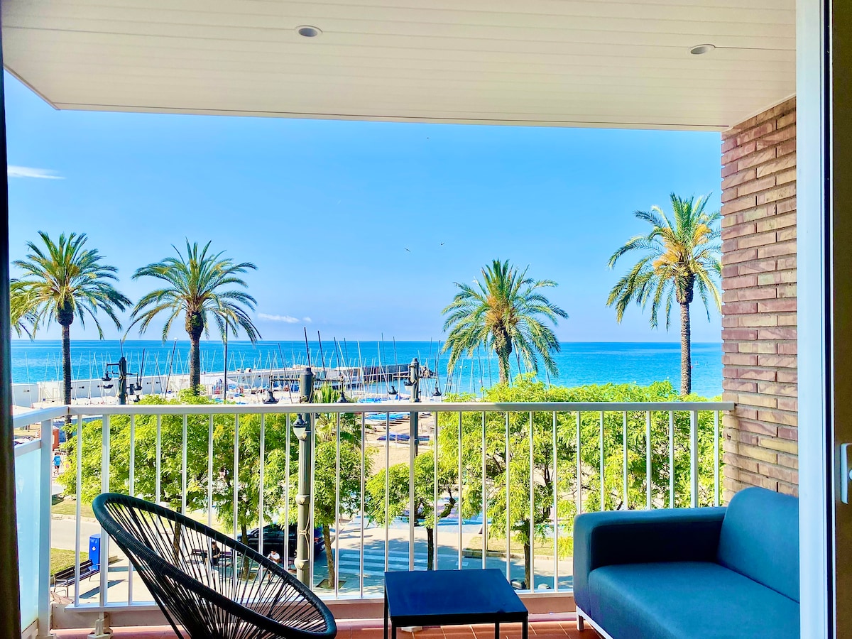 A balcony view presents a scenic line of palm trees against a clear blue sky. The sea can be seen in the distance, with soft waves lapping at the shore. A comfortable seating area is highlighted, ideal for enjoying the coastal surroundings.