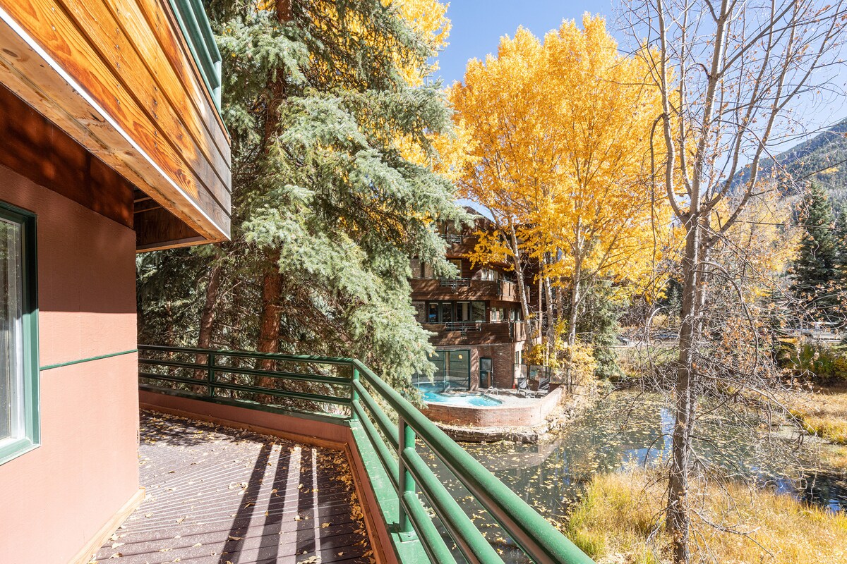 A spacious balcony features a wooden railing and overlooks a serene scene of autumn trees with vibrant yellow leaves. A hot tub is visible in the background, situated near a calm body of water, surrounded by lush greenery.