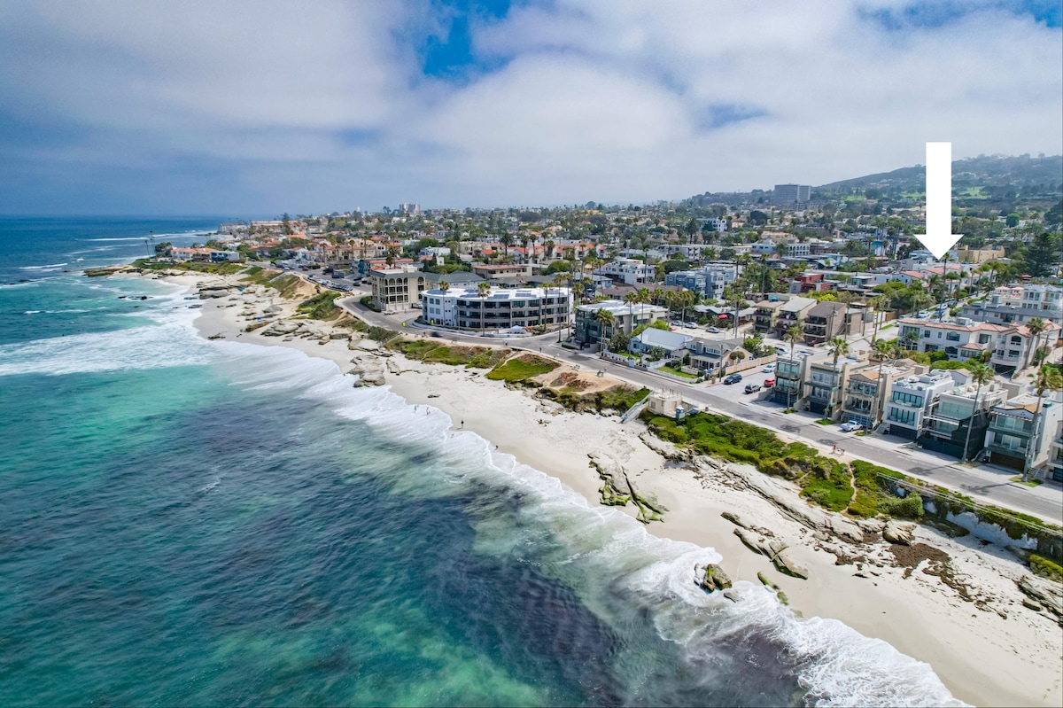 An aerial view captures the scenic coastline, showcasing gentle waves lapping against sandy shores. Nearby structures line the beach, revealing a mix of coastal homes. The vibrant blue water contrasts with the lush greenery, creating a serene coastal landscape under a partly cloudy sky.