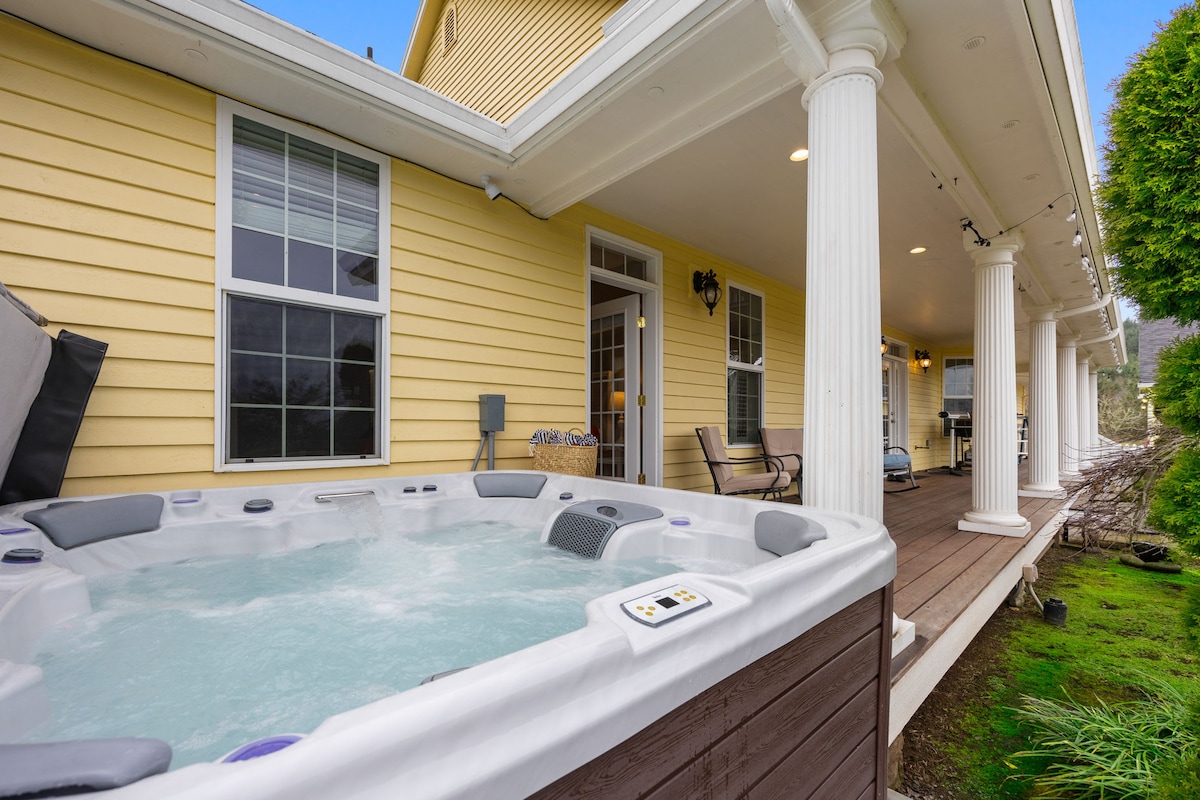 A private hot tub is situated on the patio, surrounded by a classic columned porch. Nearby, seating options are visible, providing a serene space for relaxation. Large windows showcase the peaceful exterior, illuminating the inviting atmosphere.