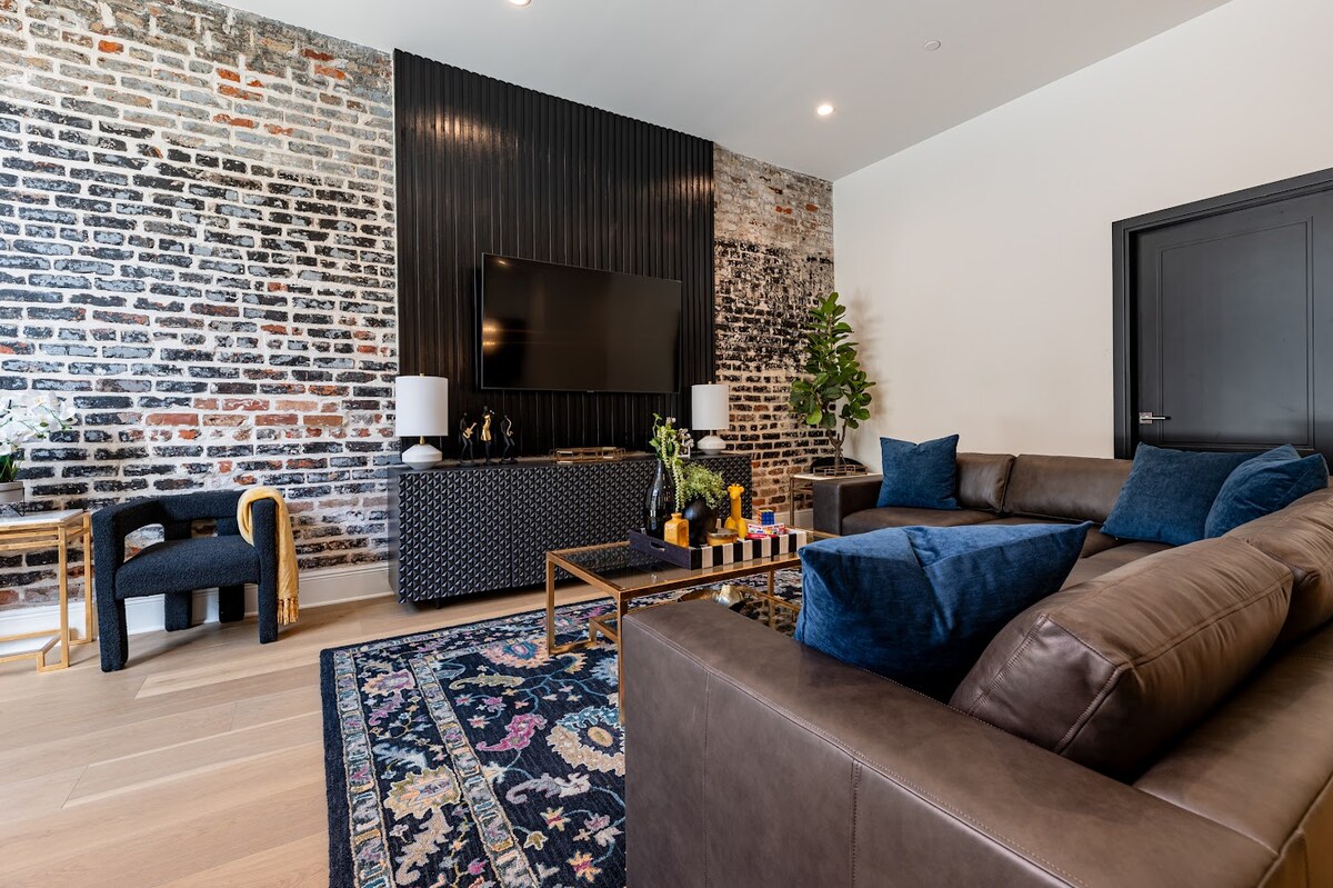The living area features a blend of modern and rustic design elements, highlighted by exposed brick walls and a sleek entertainment unit. A comfortable sectional sofa is complemented by decorative pillows, while a colorful area rug adds warmth to the space. Natural light enhances the inviting atmosphere.