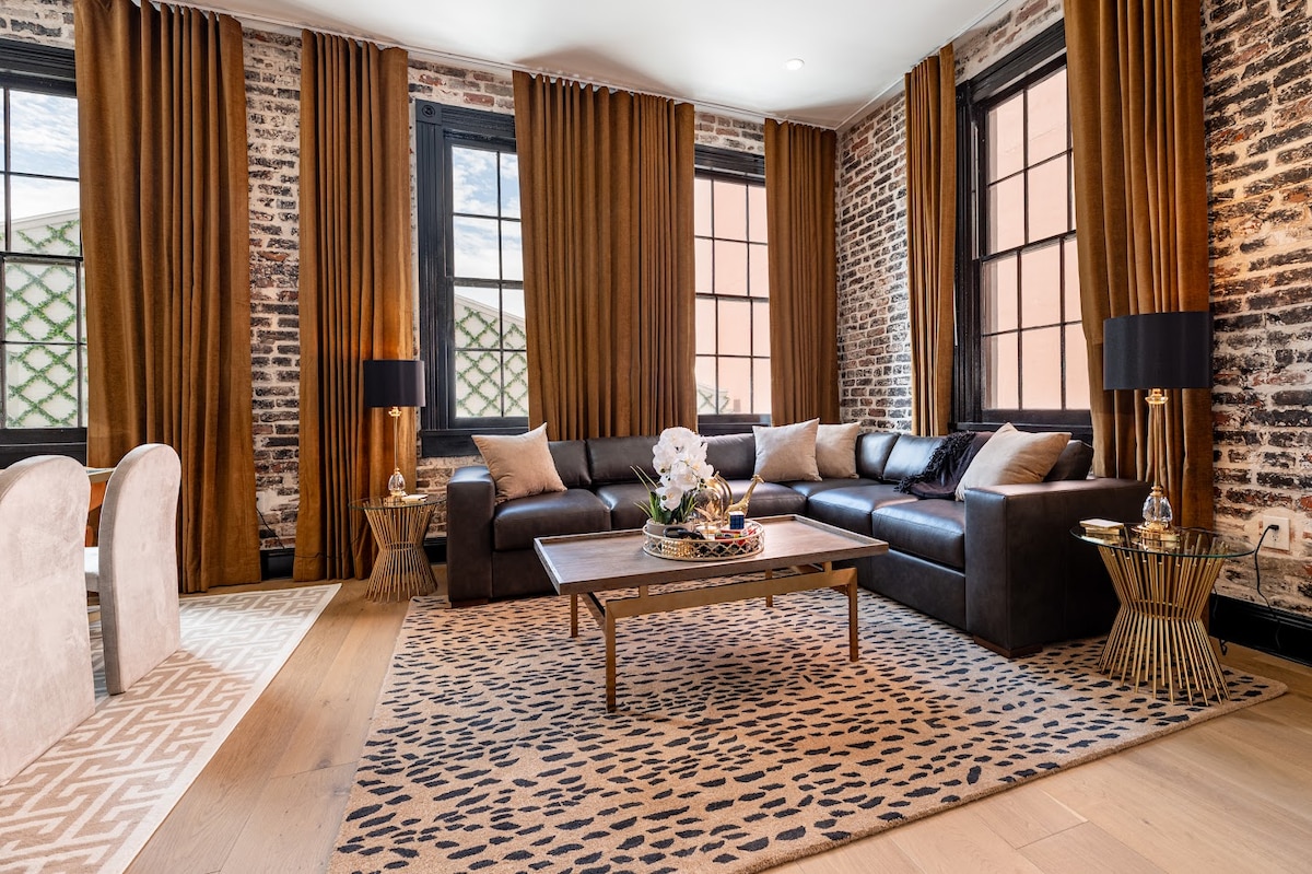 A spacious living area features a sleek, dark leather sofa arranged around a coffee table adorned with decorative items. Large windows allow natural light to illuminate the room. Rich brown curtains frame the windows, complementing the exposed brick walls and the stylish patterned rug beneath.