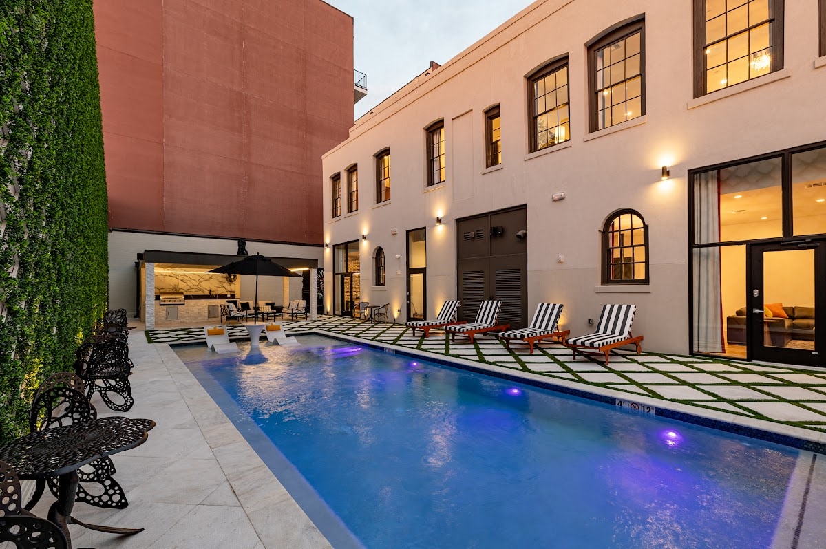 A welcoming outdoor area features a pool surrounded by lounge chairs. Shade is provided by an umbrella over seating. An outdoor kitchen area is visible in the background, framed by walls lined with greenery. Soft lighting enhances the space, creating a relaxing atmosphere.