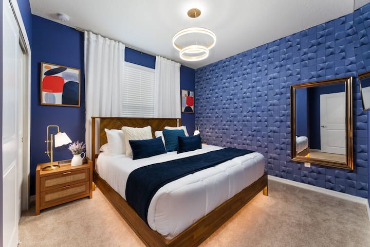 Táto štýlová spálňa má nápadnú modrú stenu, ktorá krásne kontrastuje s mäkkou neutrálnou posteľnou bielizňou a kobercom. Vychutnajte si atmosférickú žiaru moderného svietidla v kombinácii s teplým, elegantným dreveným nábytkom pre útulný pocit.