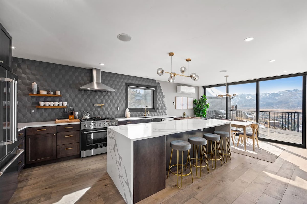 A modern kitchen with stainless steel appliances and a large marble island is showcased. Bar stools line one side of the island, while abundant natural light floods through expansive glass doors, offering panoramic mountain views. Elegant decor and fixtures enhance the space.