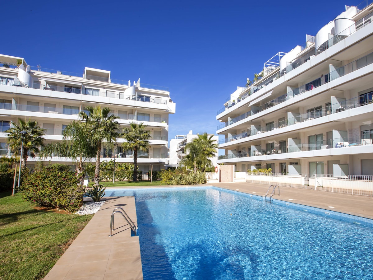 The image displays a modern apartment complex framed by well-maintained gardens with palm trees. A rectangular swimming pool is the focal point, reflecting the clear blue sky and surrounded by a spacious deck. The two adjacent buildings feature balconies adorned with greenery.