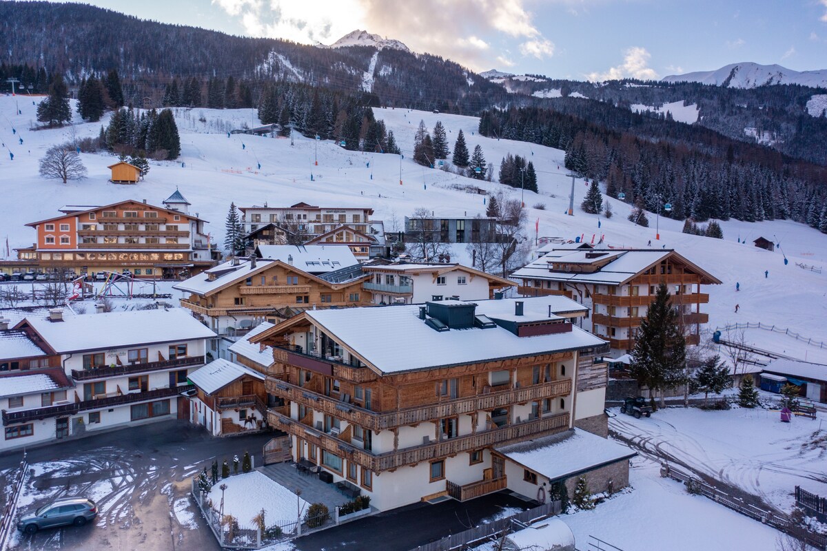 The winter landscape showcases multiple alpine-style buildings, surrounded by snow-covered terrain and evergreen trees. Slopes are visible nearby, indicating access to winter activities. The mountains in the background are partially veiled by clouds, contributing to the serene atmosphere.