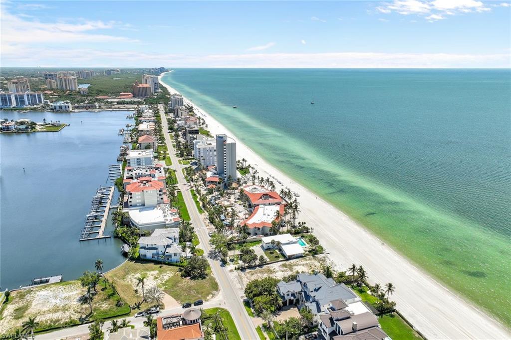 An aerial view captures the pristine coastline of Vanderbilt Beach, featuring smooth sandy shores and tranquil turquoise waters. Lush palm trees line the beach, while elegant condominiums and homes are visible alongside a calm waterway, creating a serene coastal atmosphere.
