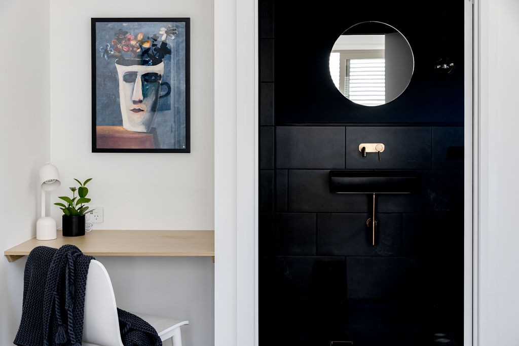 A sleek bathroom area features black tiled walls and modern fixtures, including a circular mirror and a stylish black sink. A small desk with a light and a potted plant adds warmth to the space, while a cozy throw is draped over a white chair.