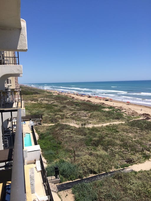A panoramic view from a private balcony showcases the sandy beach stretching along the coastline, with gentle waves lapping at the shore. Beach umbrellas dot the shoreline, and a pool area is visible below, surrounded by vegetation.