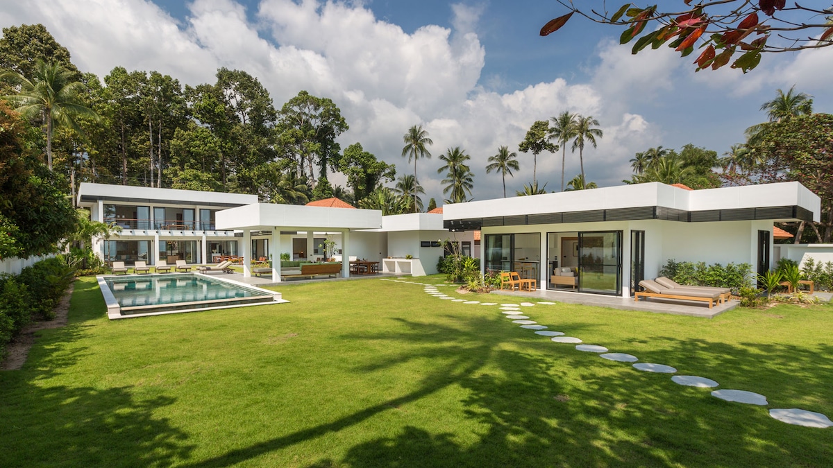 A modern villa is depicted, featuring a spacious green lawn that leads to a swimming pool. The structure comprises multiple segments with large glass doors, providing a connection between indoor and outdoor spaces. Palm trees and tropical foliage surround the property.