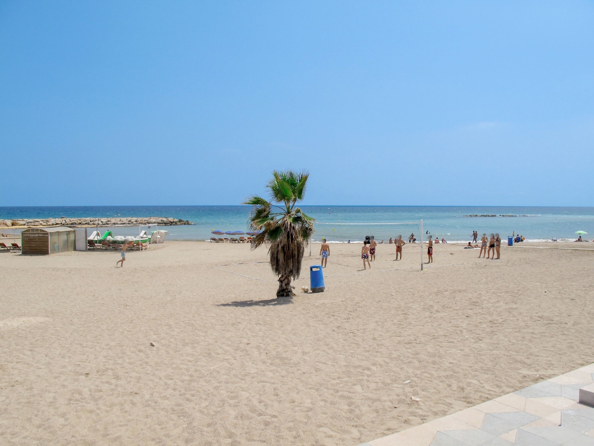 A sandy beach stretches into the distance, with gentle waves lapping at the shore. A palm tree stands in the foreground, while beachgoers are seen enjoying the sun. Umbrellas and beach chairs are positioned along the coastline, creating a relaxed atmosphere.