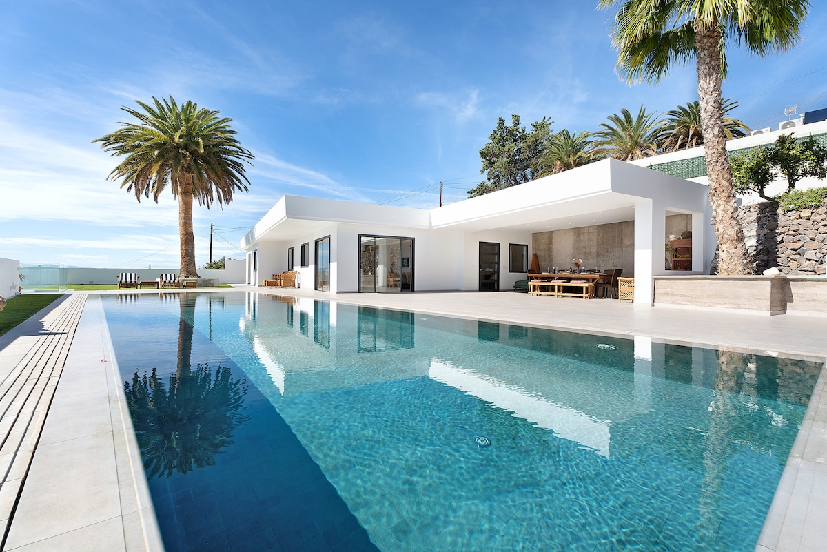 An expansive outdoor area showcases a pristine pool reflecting the clear sky and surrounded by palm trees. The modern structure features large glass doors that offer views into the bright living spaces. A dining table, positioned beside the pool, invites outdoor gatherings.