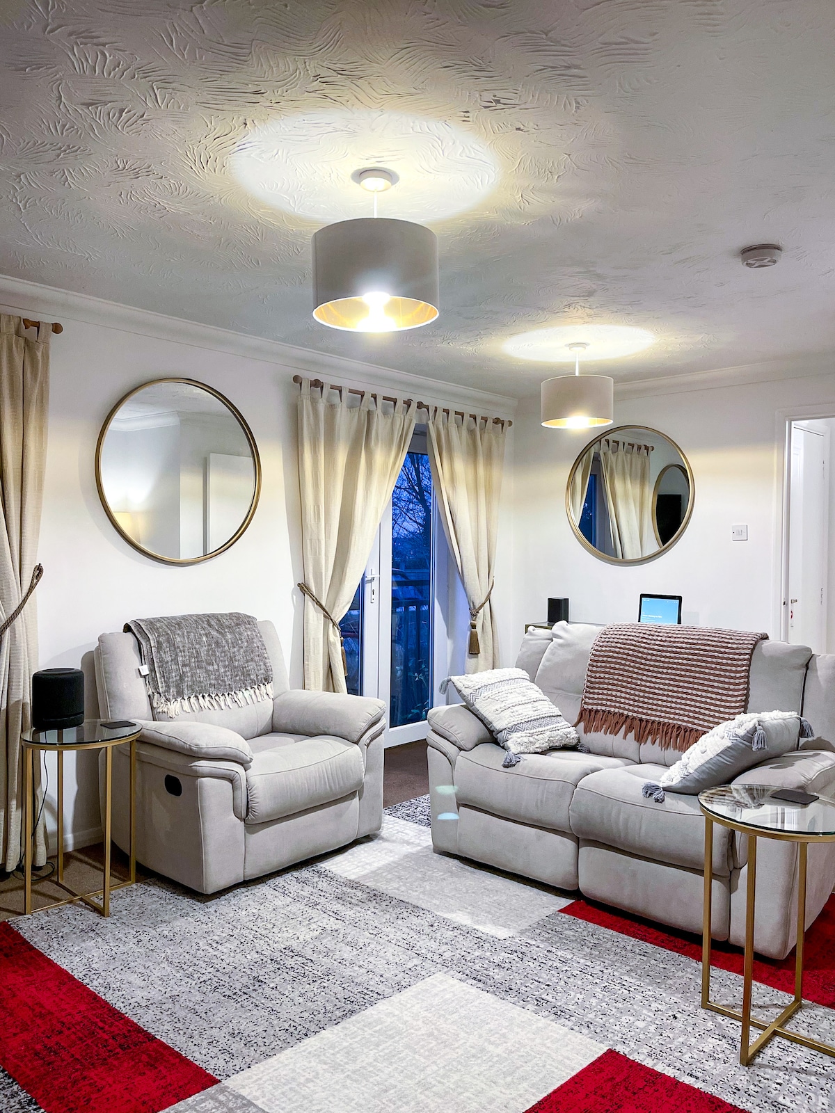 A cozy living area features two light-colored reclining chairs and a sofa, accented by decorative throw blankets. Round mirrors are mounted on the walls, and natural light enters through a sliding door, which is partially covered by curtains. A patterned rug adds visual interest to the space.