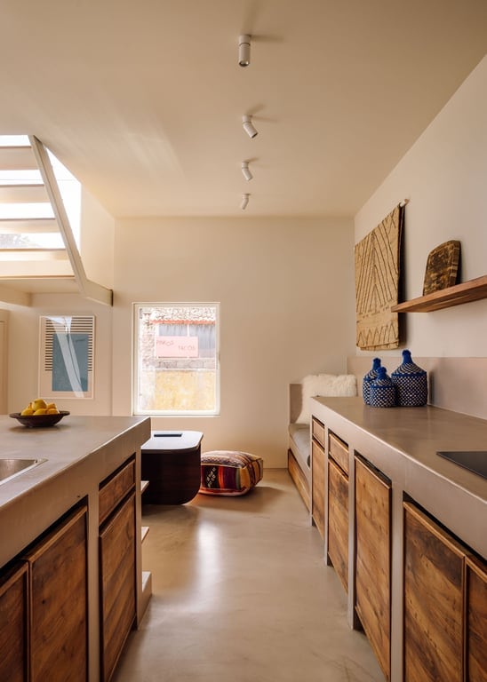 A minimalist kitchen space is presented with natural wood cabinetry and a light-colored countertop. A small seating area is visible, featuring a colorful pouf. Natural light filters through a window, enhancing the calm atmosphere. Decorative items are displayed on the walls.