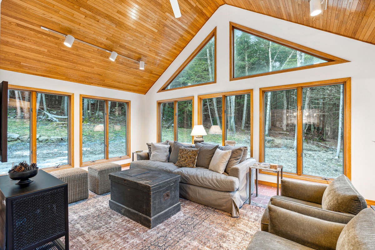 A bright living area is enveloped with natural light from large windows framed in wood, showcasing views of the surrounding greenery. Cozy seating is arranged around a rustic coffee table, creating an inviting space for relaxation. The warm wooden ceiling adds to the room's comfortable ambiance.