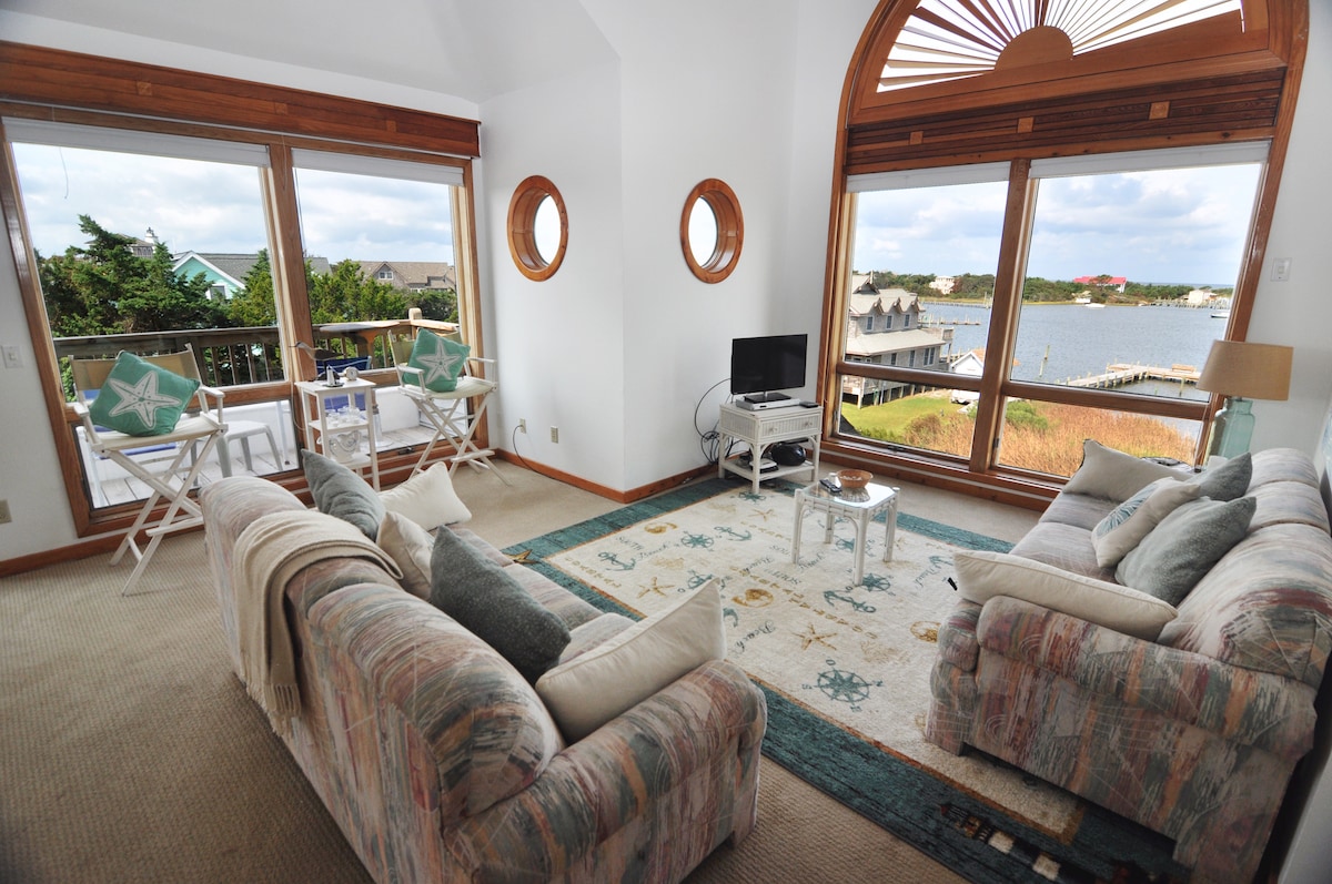A spacious living area features large windows showcasing views of Silver Lake Harbor. Comfortable sofas are arranged around a central decorative rug. Two chairs are positioned near the windows, providing a cozy spot for relaxation. A television is visible in one corner of the room.